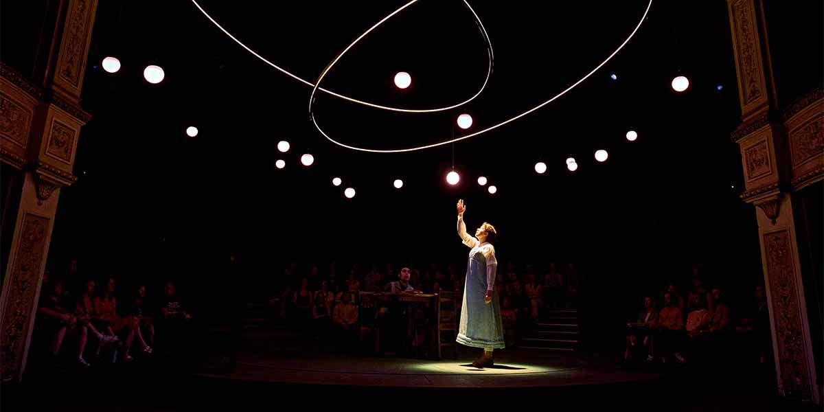 Isis Hainsworth as Thomasina Coverly reaches towards suspended lights, surrounded by an audience in darkness.