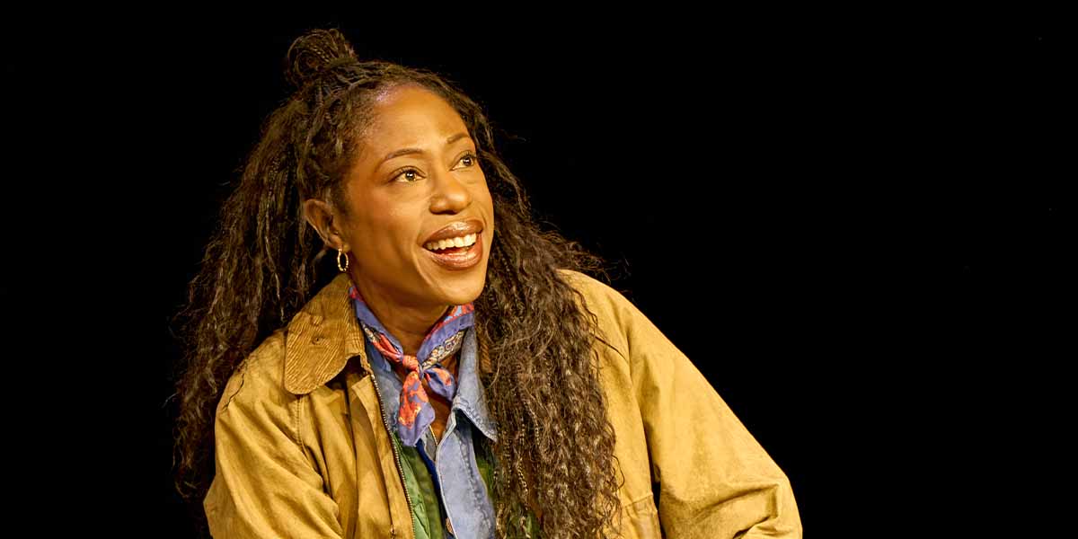 Nikki Amuka-Bird as Hannah Jarvis smiles, wearing a brown jacket and blue scarf against a black background.