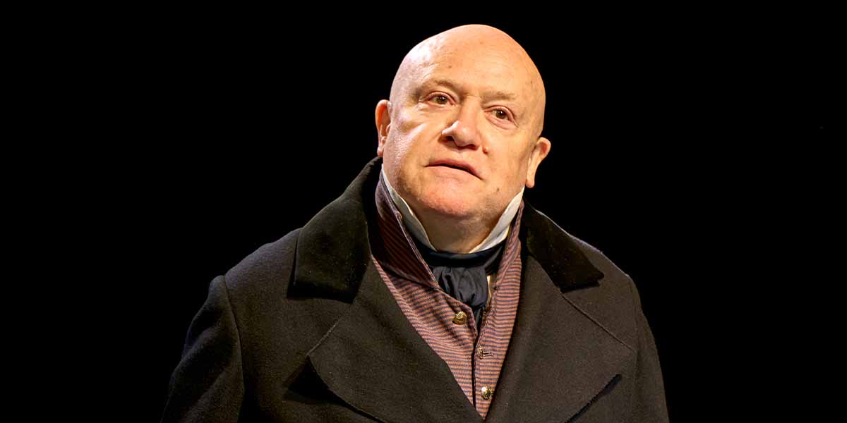 Tim Frances as Jellaby, wearing a dark coat and patterned shirt, gazes thoughtfully against a black background.