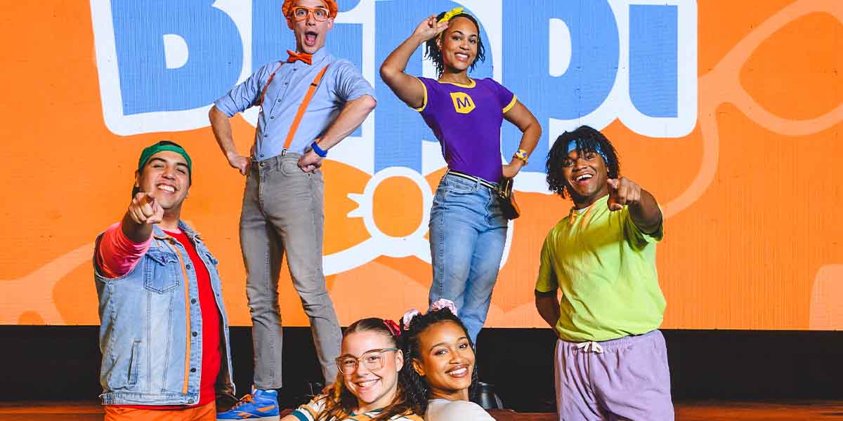Six performers pose playfully on stage, with a bright orange backdrop featuring "Blippi" text.