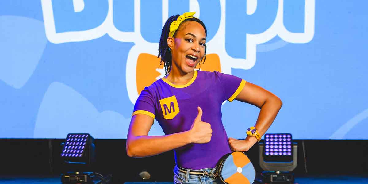 A woman in a purple shirt with yellow accents smiles and gives a thumbs-up against a colourful backdrop.