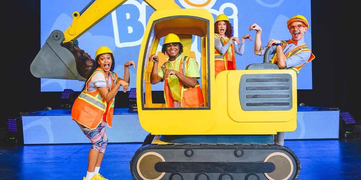 Four performers in safety vests and hard hats pose playfully around a large yellow excavator on stage.