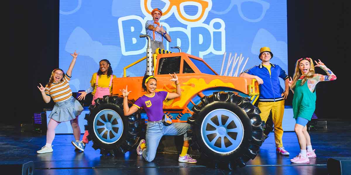 Six performers pose energetically around a large orange toy truck, with a colourful backdrop displaying "Blippi.