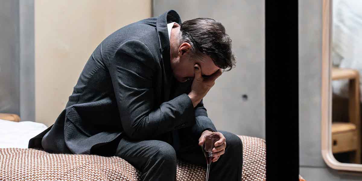 David Morrissey sits on a bed, head in hand, wearing a dark suit, conveying distress.