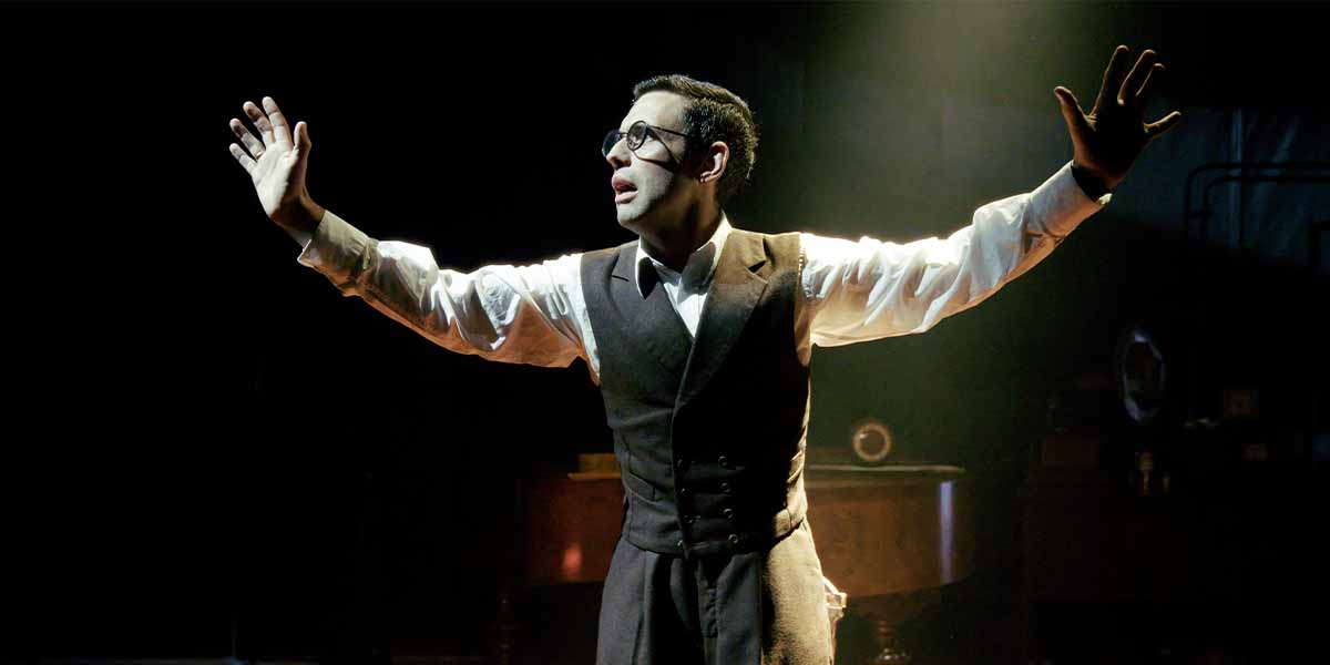 A performer in a suit and glasses gestures dramatically under spotlight, with a dark background.