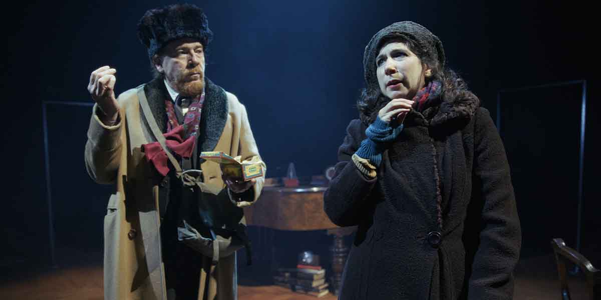 A man in a coat and hat gestures while a woman in a dark coat looks thoughtfully.