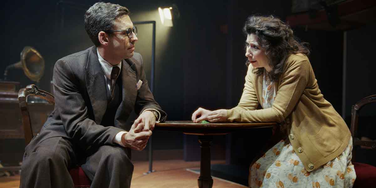 Two actors seated at a table, engaged in conversation. One wears a suit, the other a cardigan.