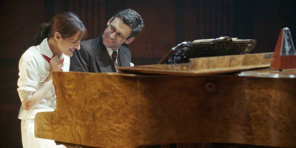 A woman in a white outfit and a man in a suit smile at each other by a piano.