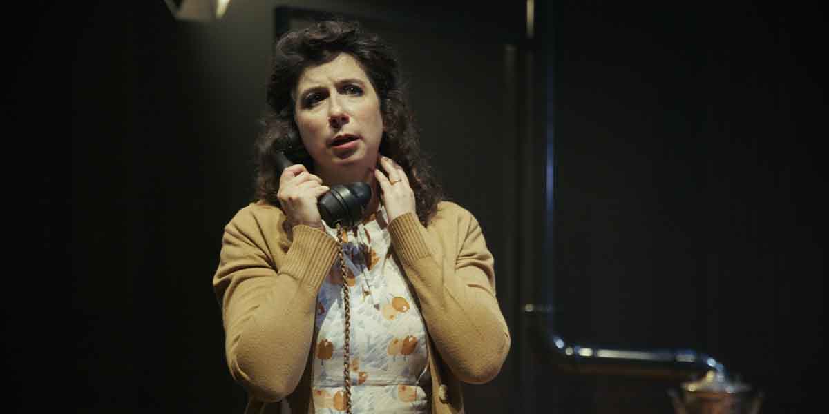 A woman with dark hair holds a vintage telephone, looking concerned, wearing a patterned dress and cardigan.