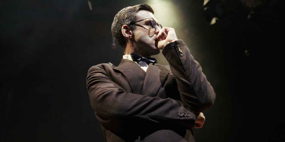A man in a dark suit and bow tie poses thoughtfully, illuminated by stage lights in a theatre.