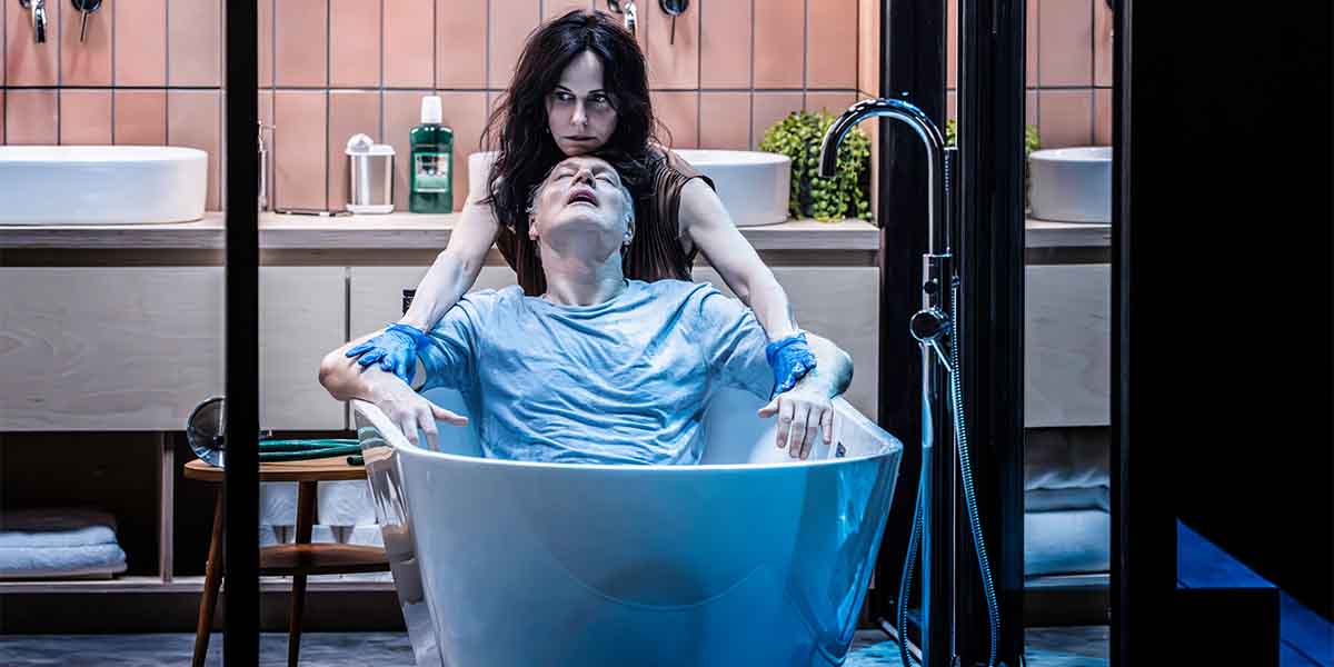 Mary-Louise Parker leans over David Morrissey, who is reclining in a white bathtub, in a modern bathroom.