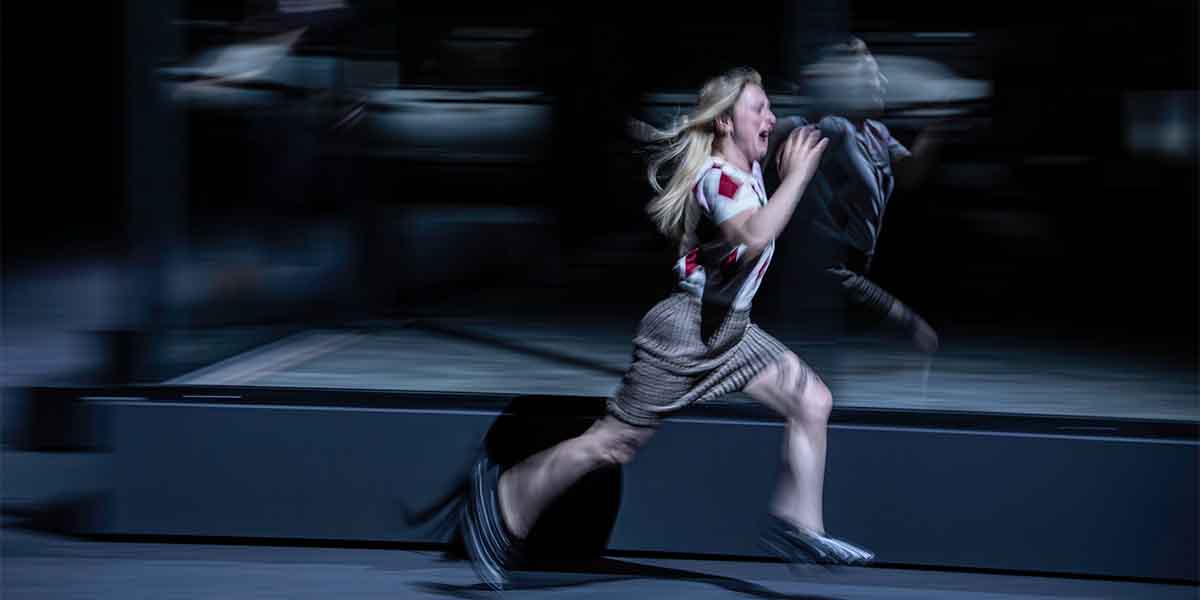 A woman with long hair runs energetically across a dark stage, wearing a striped shirt and skirt.