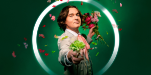 A person dressed as as Oscar Wilde, wearing a dark suit and bow tie, seated on a chair, smiling and holding flowers.