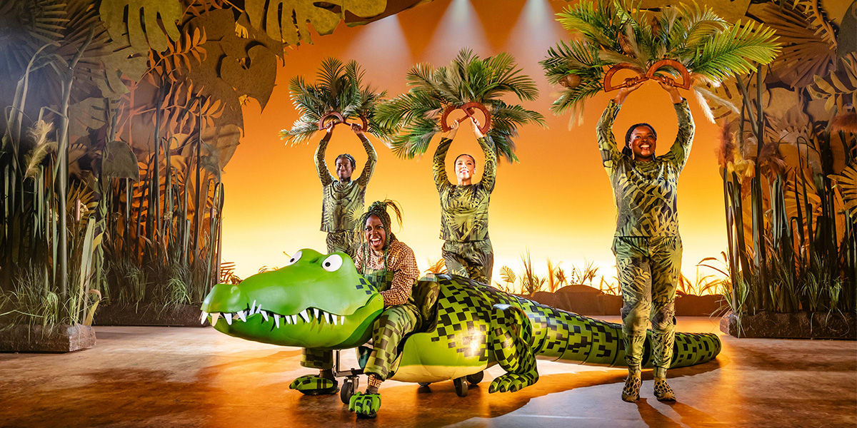 A crocodile puppet with vibrant green scales and yellow eyes is centre stage in a theatrical setting.