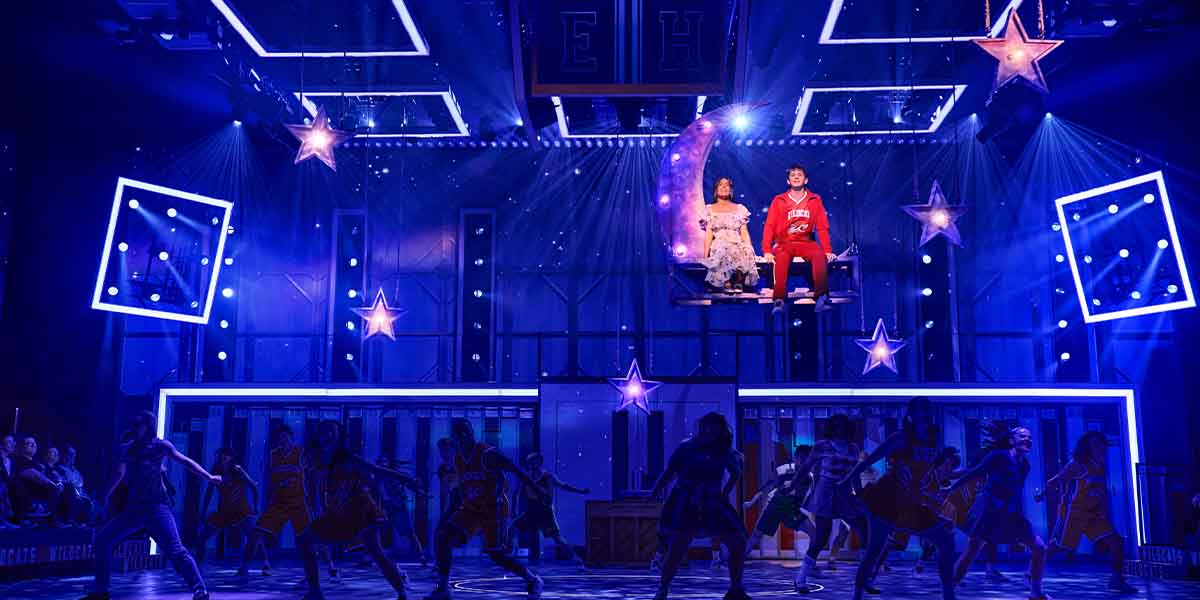 Leonor Correia as Gabriella Montez and Tobias Turley as Troy Bolton perform on stage, surrounded by dancers.
