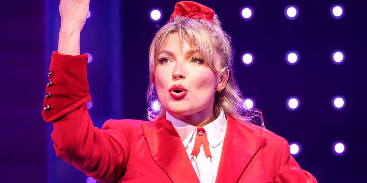 Liberty Stottor performs passionately in a red outfit, gesturing with one hand against a lit backdrop.
