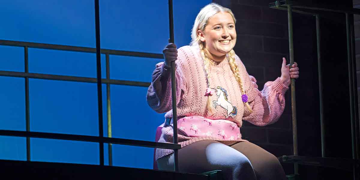 Sophie Manners smiles while sitting on a window ledge, wearing a pink sweater with a unicorn design.