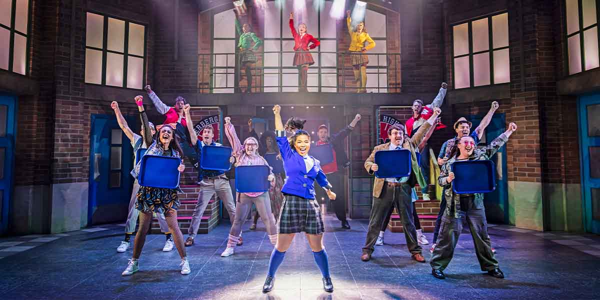 A diverse cast performs energetically on stage, holding blue trays, with vibrant costumes and dramatic lighting.