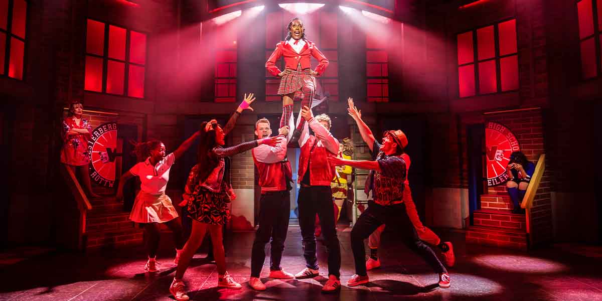 A performer in a red outfit is elevated by cast members, surrounded by dramatic lighting and vibrant costumes.