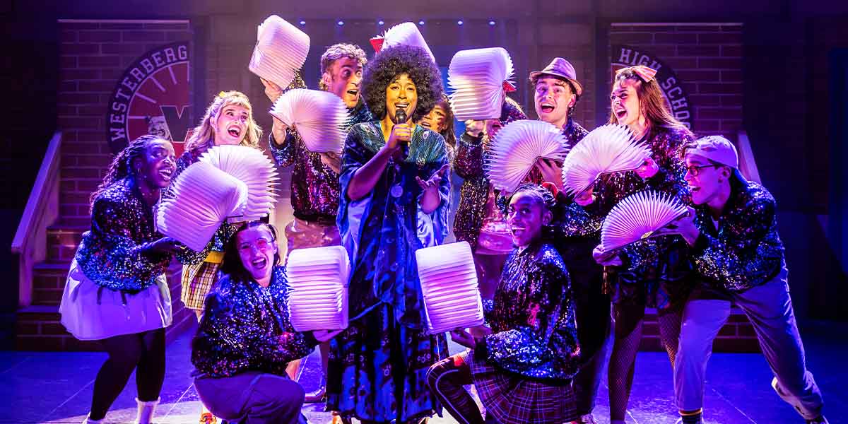 The cast of Heathers The Musical poses together, wearing vibrant costumes, with a colourful stage backdrop.