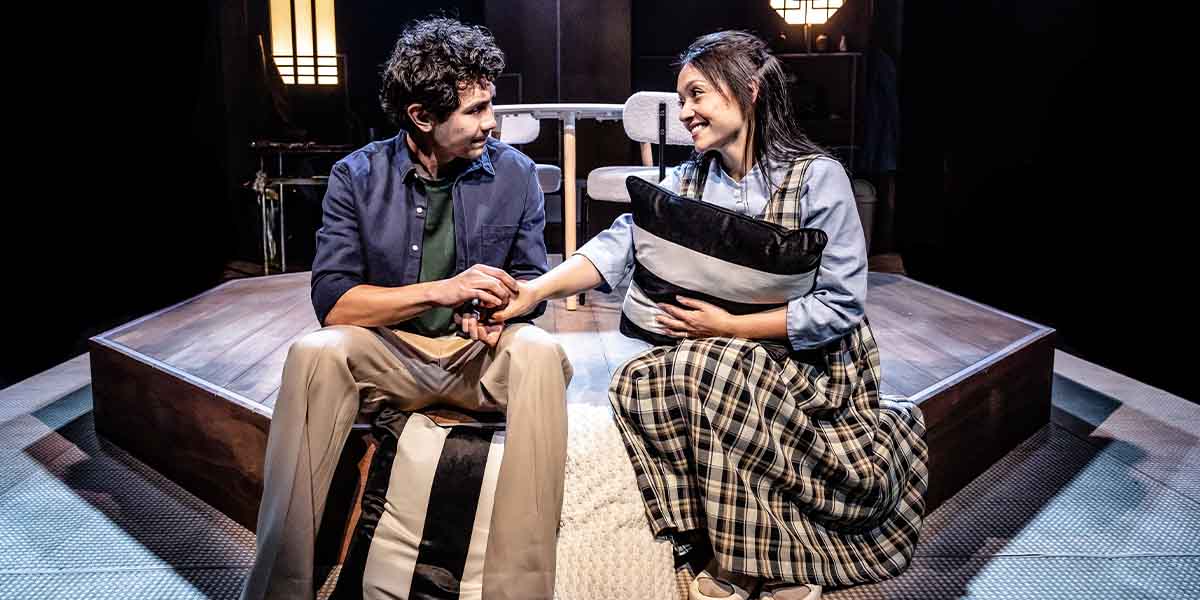 Two actors sit on stage, holding hands and smiling, surrounded by dark lighting and wooden set pieces.