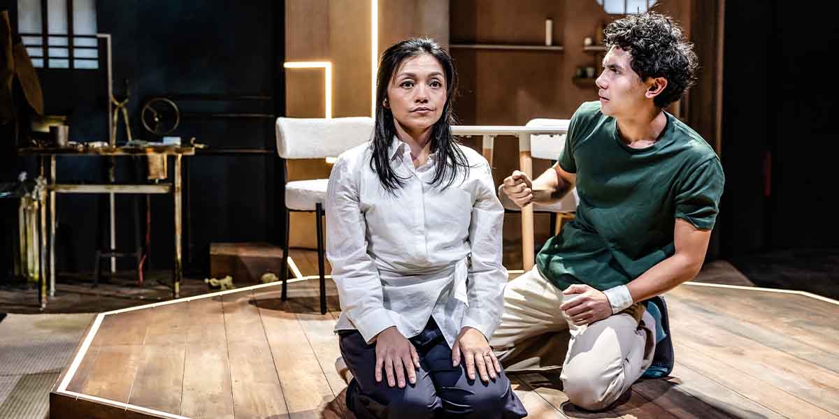 A woman in a white blouse sits on a stage, while a man in a green shirt gestures towards her.