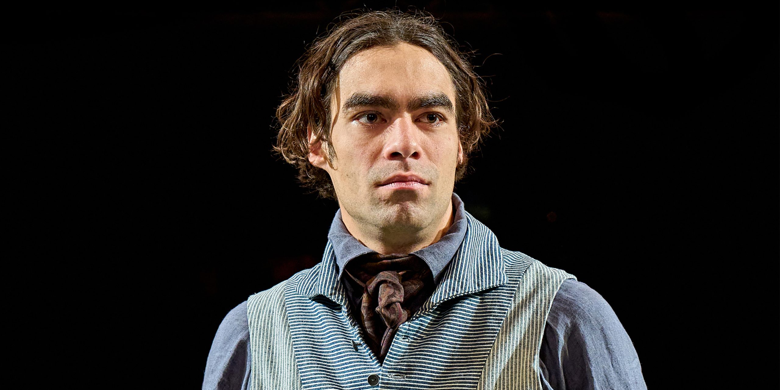 Seamus Dillane speaking into a microphone, wearing a dark jacket, with a blurred audience in the background.
