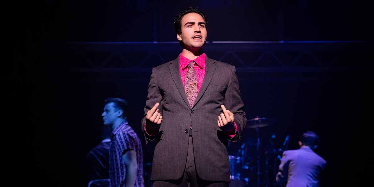 Carlo Boumouglbay as Tommy DeVito performs on stage, wearing a grey suit and pink shirt.