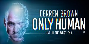 Derren Brown performs on stage, illuminated by bright lights, with a focused expression and gesturing hands.