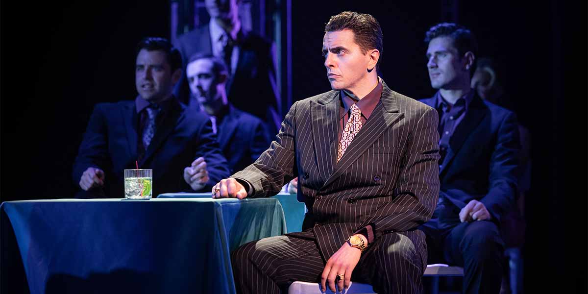Fed Zanni as Gyp DeCarlo sits at a table, wearing a pinstriped suit, with other people in shadows.