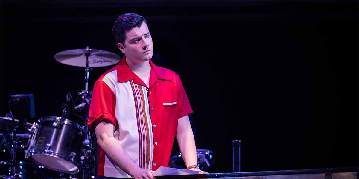 Lewis Kennedy as Nick Massi, wearing a red and white shirt, looks pensively at a script.