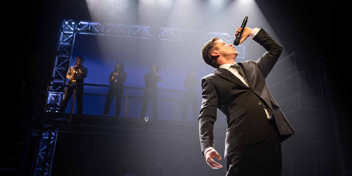Luke Baker as Frankie Valli sings passionately on stage, dressed in a black suit with a microphone.