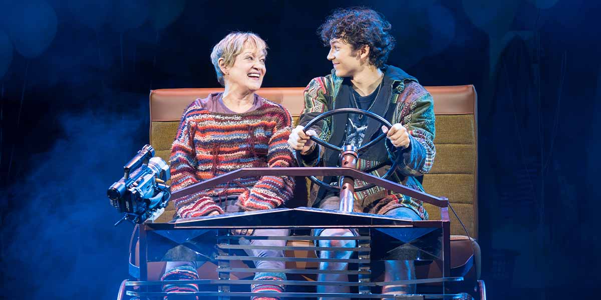 Maria Friedman as Kimberly Levaco and Gilli Jones as Seth Weetis smile while seated in a vehicle.