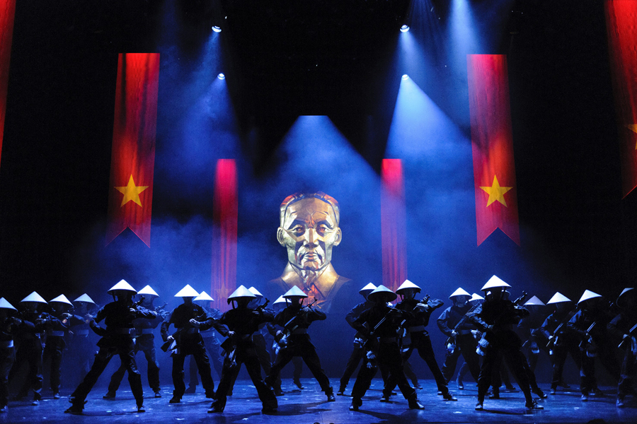 A dramatic stage scene from Miss Saigon, featuring performers in traditional hats, with a large portrait backdrop.