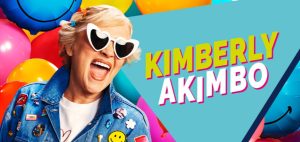 Kimberly Akimbo stands centre stage, smiling, wearing a floral dress, with a vibrant backdrop of lights.
