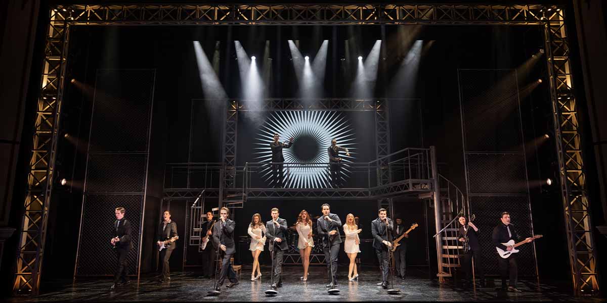 The cast of Jersey Boys performs on stage, dressed in black and white, with dramatic lighting.