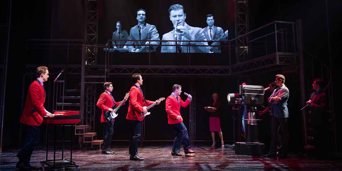 The cast of Jersey Boys performs on stage, wearing red jackets, with a large screen backdrop.