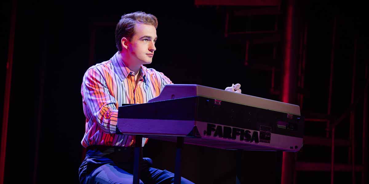 Toby Miles as Bob Gaudio sits at a Farfisa keyboard, wearing a colourful striped shirt, focused.