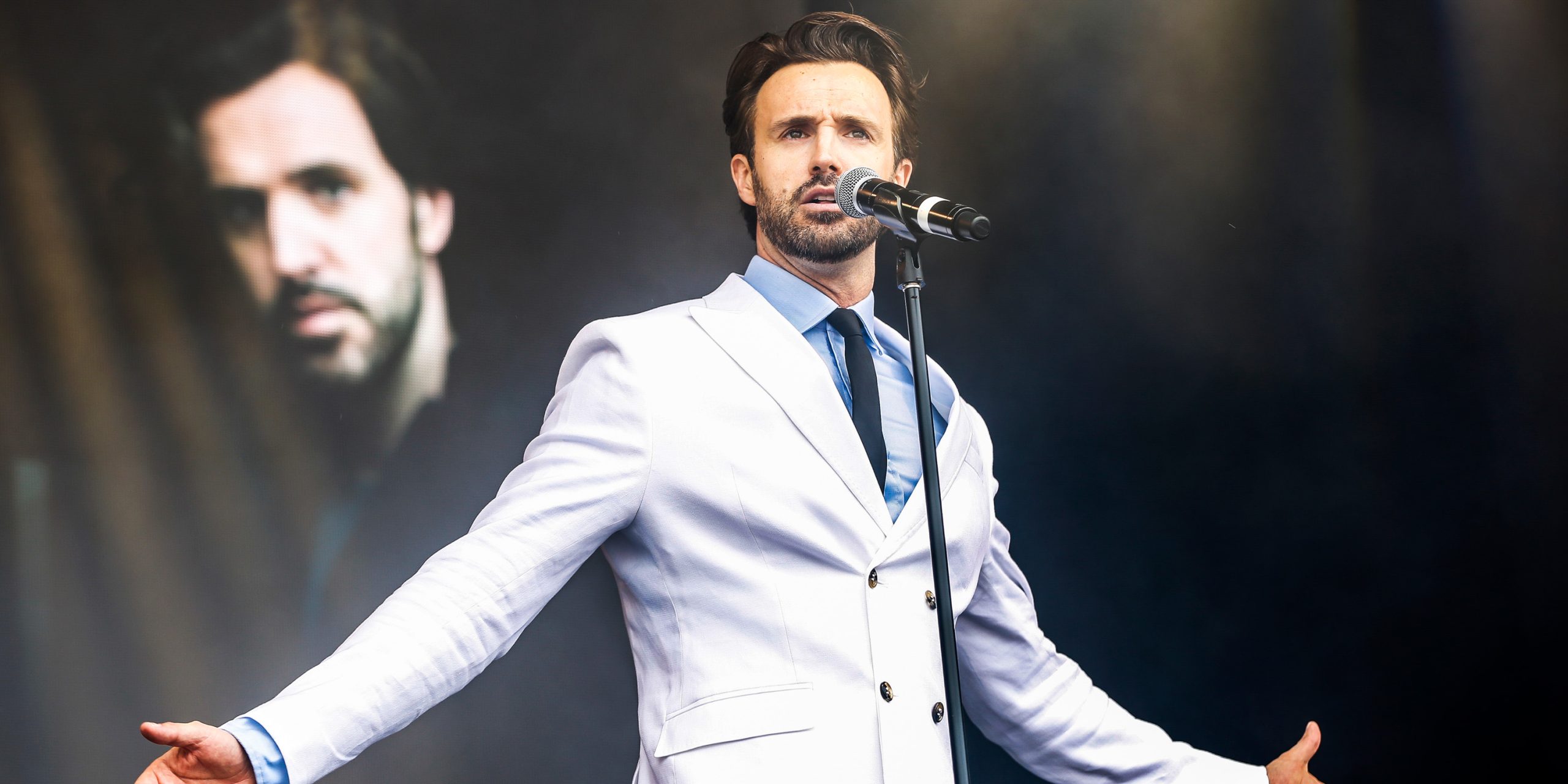 Glenn Adamson performs on stage in a white suit, microphone in hand, with a dramatic expression.