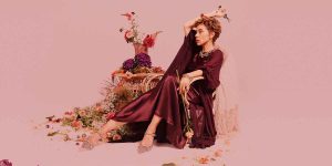 A woman in a flowing burgundy dress poses elegantly on a chair surrounded by colourful flowers.