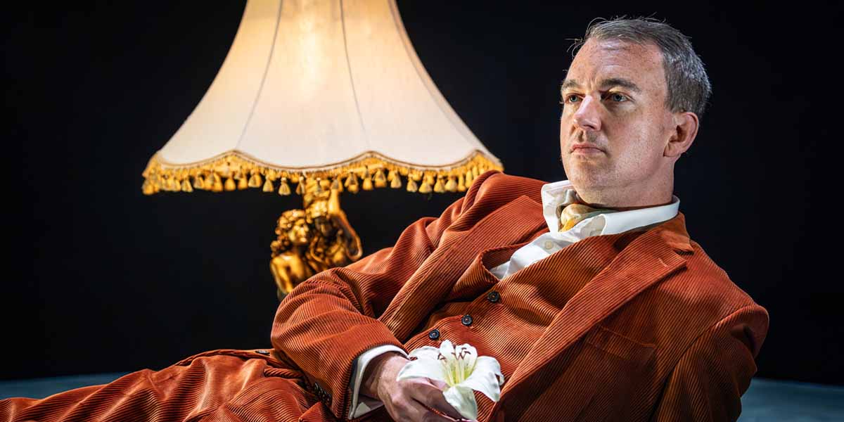 Alastair Whatley reclines in an orange suit, holding a white flower, beside a decorative lamp.
