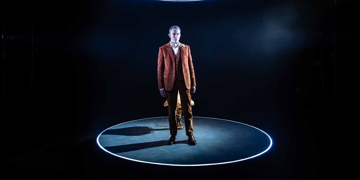 Alastair Whatley stands in a spotlight, wearing an orange suit, against a dark background.