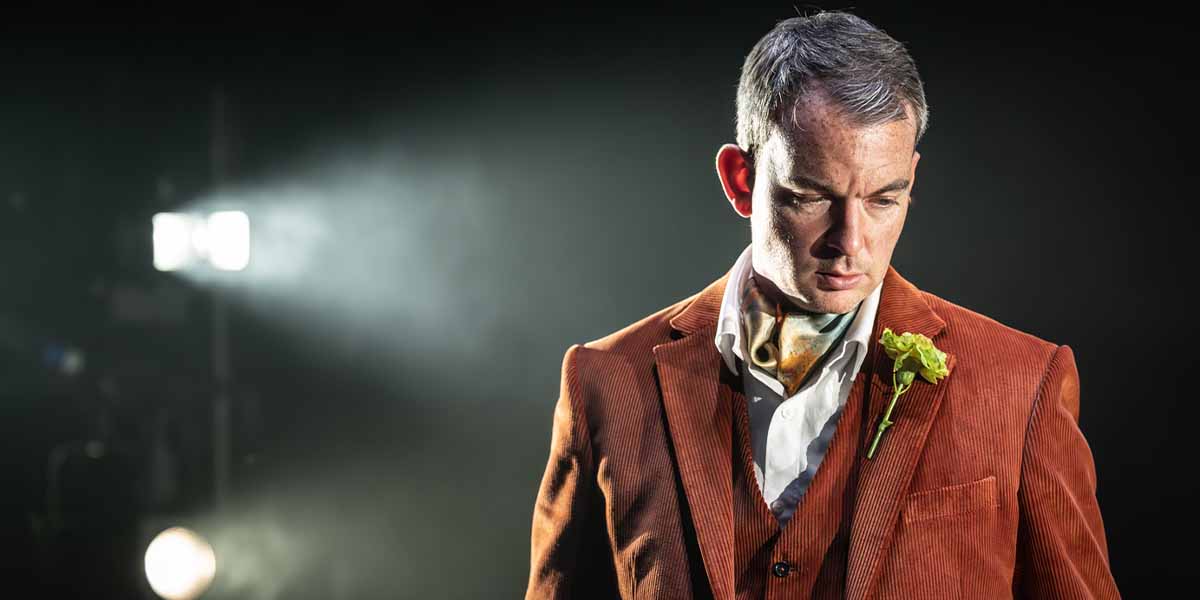 Alastair Whatley, wearing an orange corduroy suit, gazes downwards with a flower pinned to his lapel.