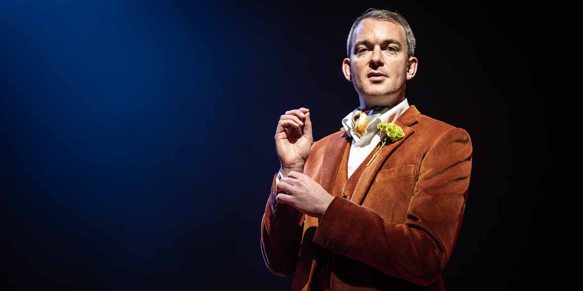 Alastair Whatley stands in a spotlight, wearing a brown corduroy suit and a white shirt with a flower.