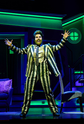 A performer in a striped suit gestures dramatically on stage, with a vibrant green and blue backdrop.