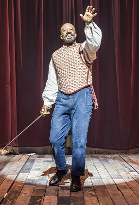 A performer in period costume gestures dramatically on stage, with a red curtain backdrop and wooden floor.